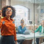 Mulher negra escrevendo em parede de vidro enquanto colabora com equipes