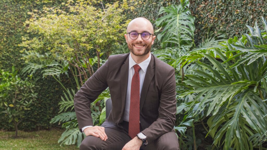 Homem sorridente, de terno e gravata, sentado em ambiente externo com vegetação ao fundo 